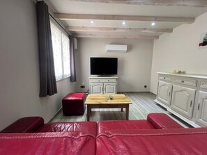 Ein Wohnzimmer mit einem roten Ledersofa, einem Holztisch, einem Fernseher auf einem weißen Schrank und einem Fenster mit Vorhängen.