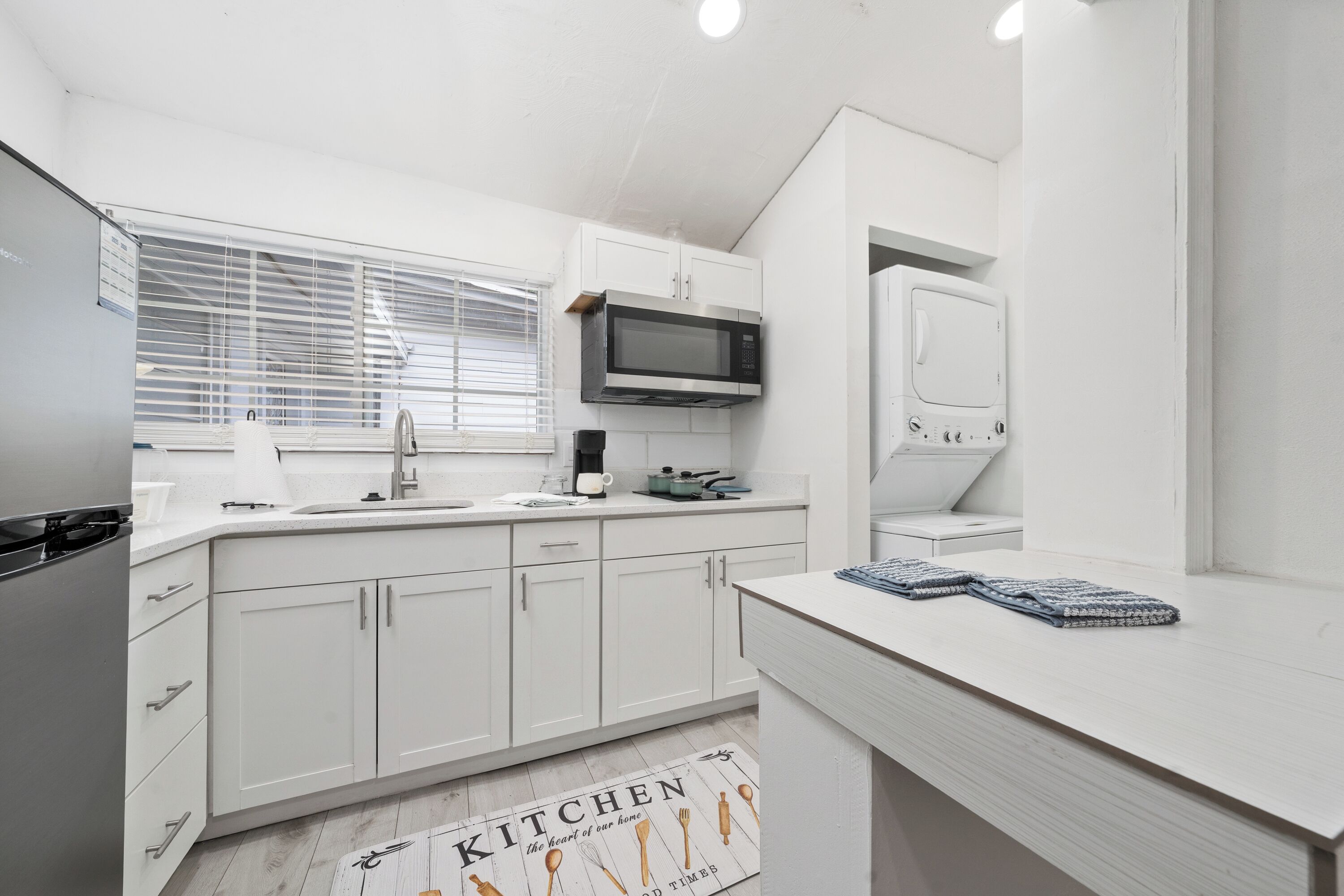 Una cocina moderna con armarios blancos, microondas, hervidor y ventana con persianas.