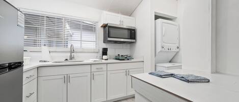 Una cocina moderna con armarios blancos, microondas, hervidor y ventana con persianas.