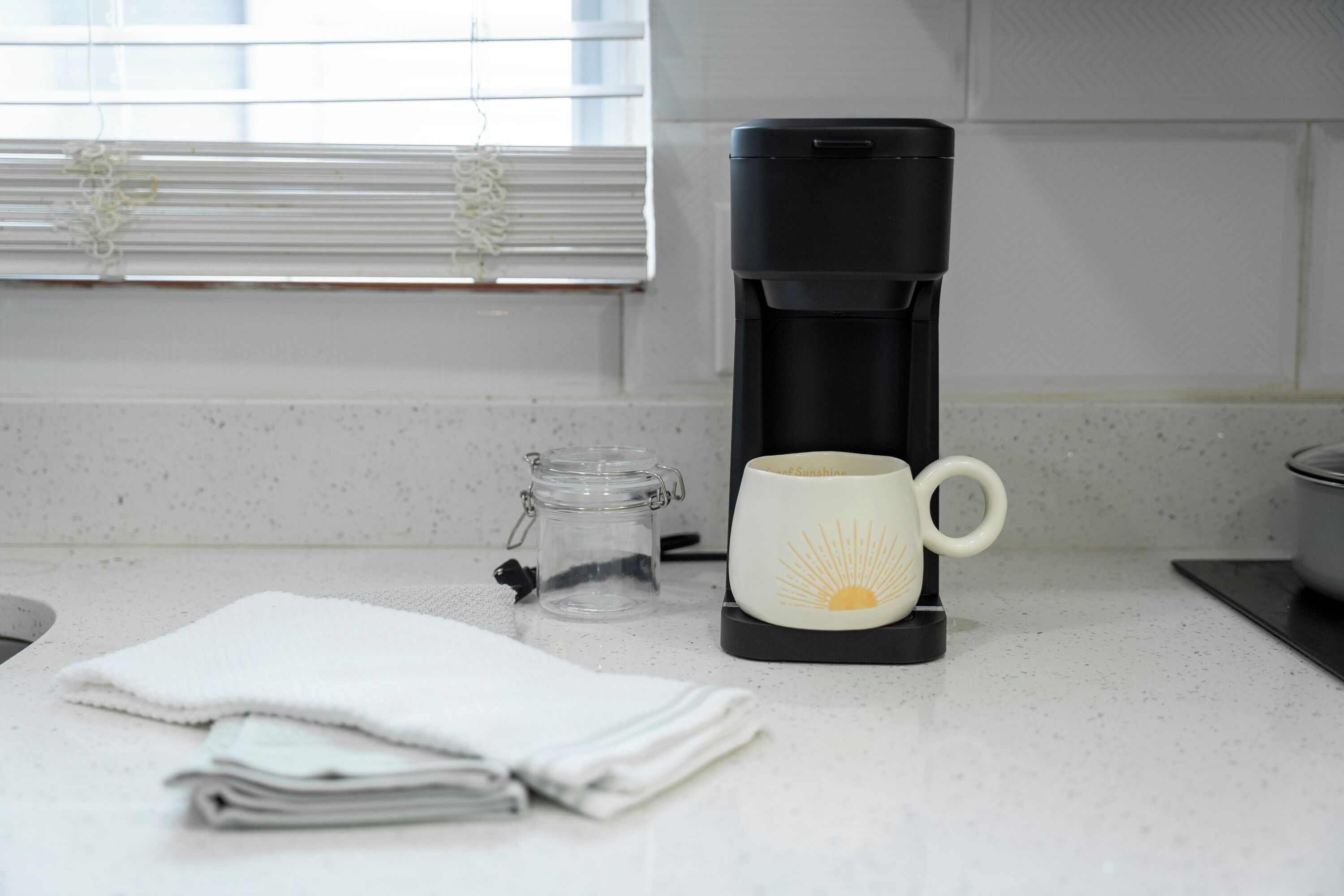 A kitchen counter with a coffee maker, a mug, a glass jar, and a folded towel.