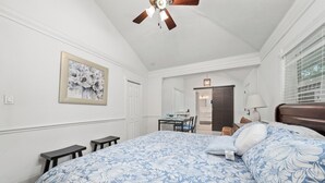 Un dormitorio con una cama grande, un ventilador de techo, una mesa de comedor con sillas y un cuadro con flores en la pared.