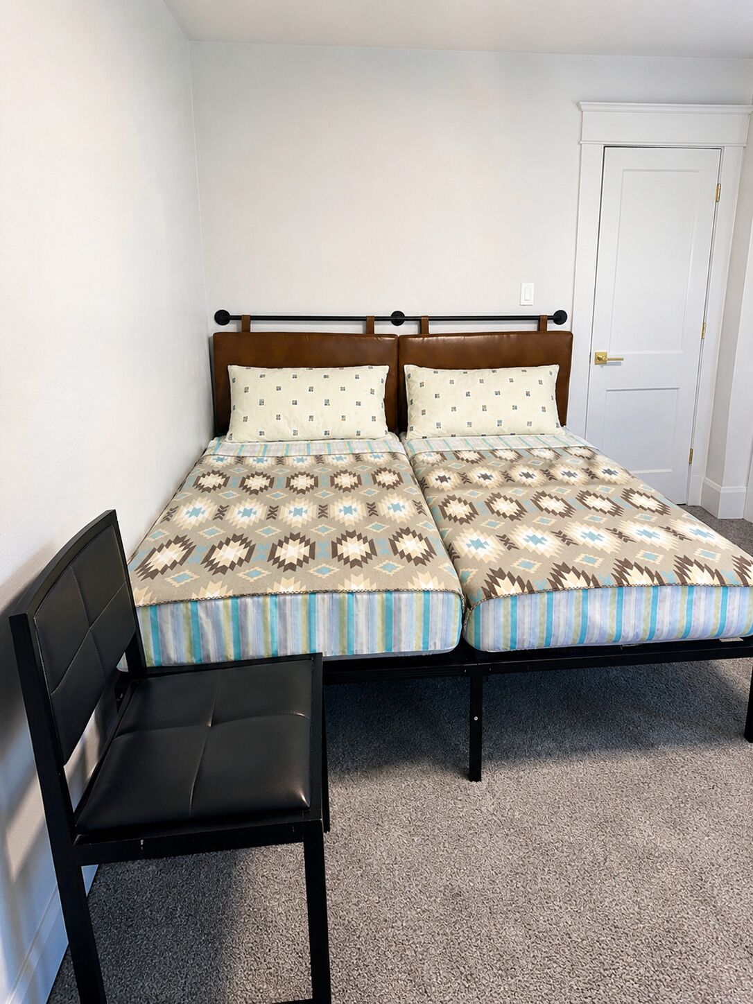 Ein Doppelbett mit gemusterten Betttextilien, ein Holzkopfschild, ein schwarzer Stuhl und eine weiße Tür.