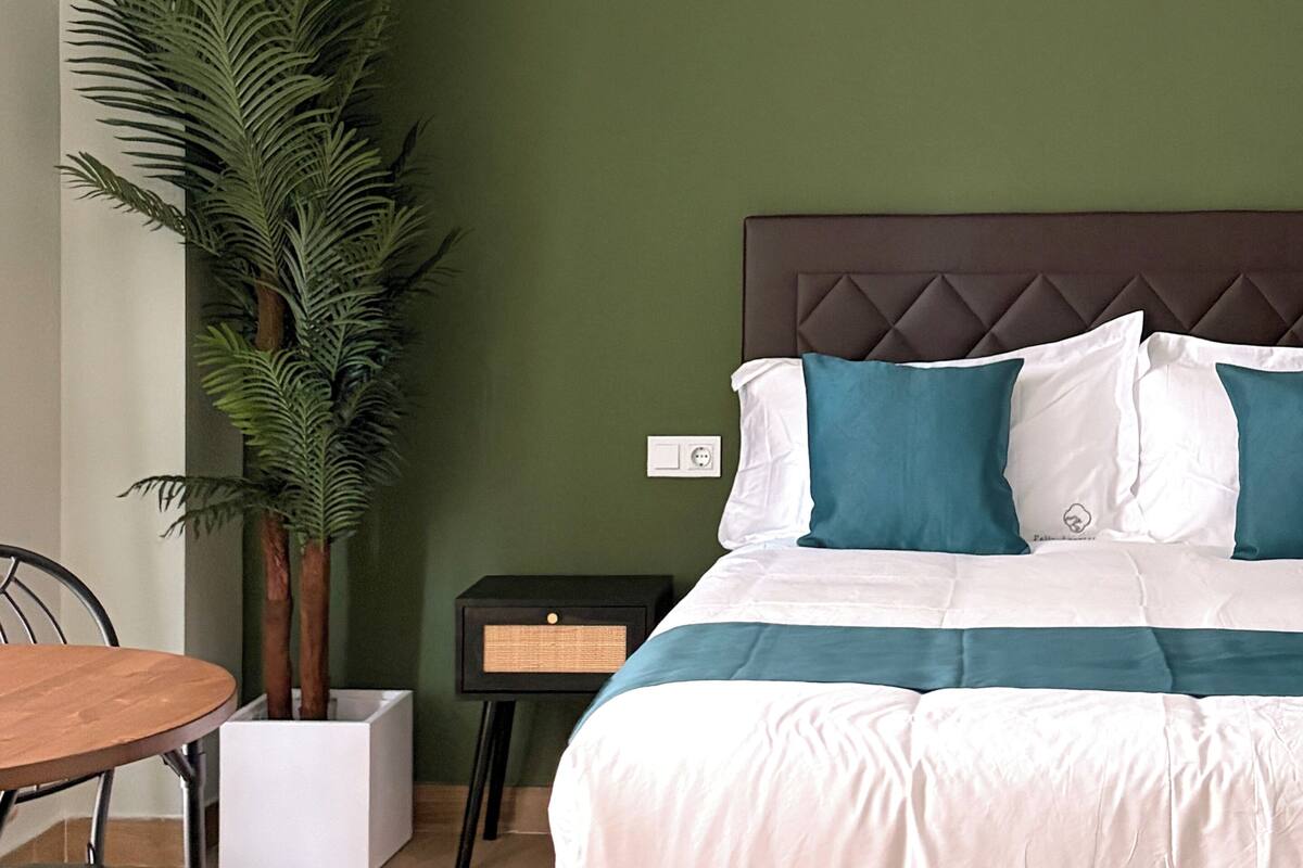 A bedroom with a bed, a nightstand, a chair, a table, and a potted plant.