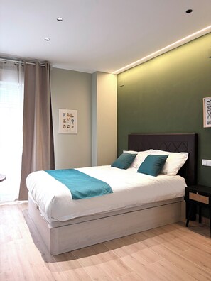 A neatly made bed with a white and teal bedspread, a dark headboard, and a green wall with a framed picture. - Feliz Apartments (Madrid)