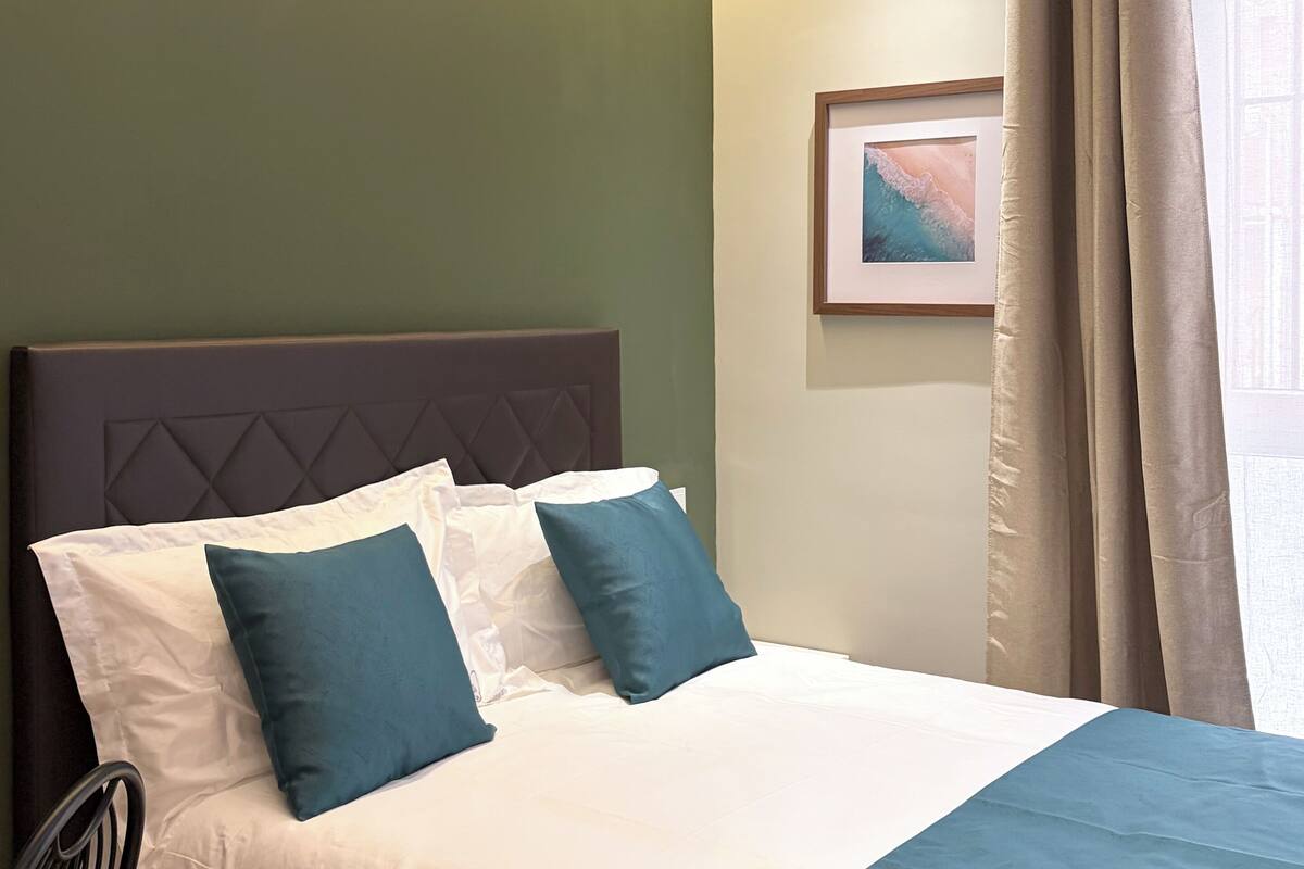 A hotel room with a bed, a headboard, a framed picture on the wall, and a window with curtains.