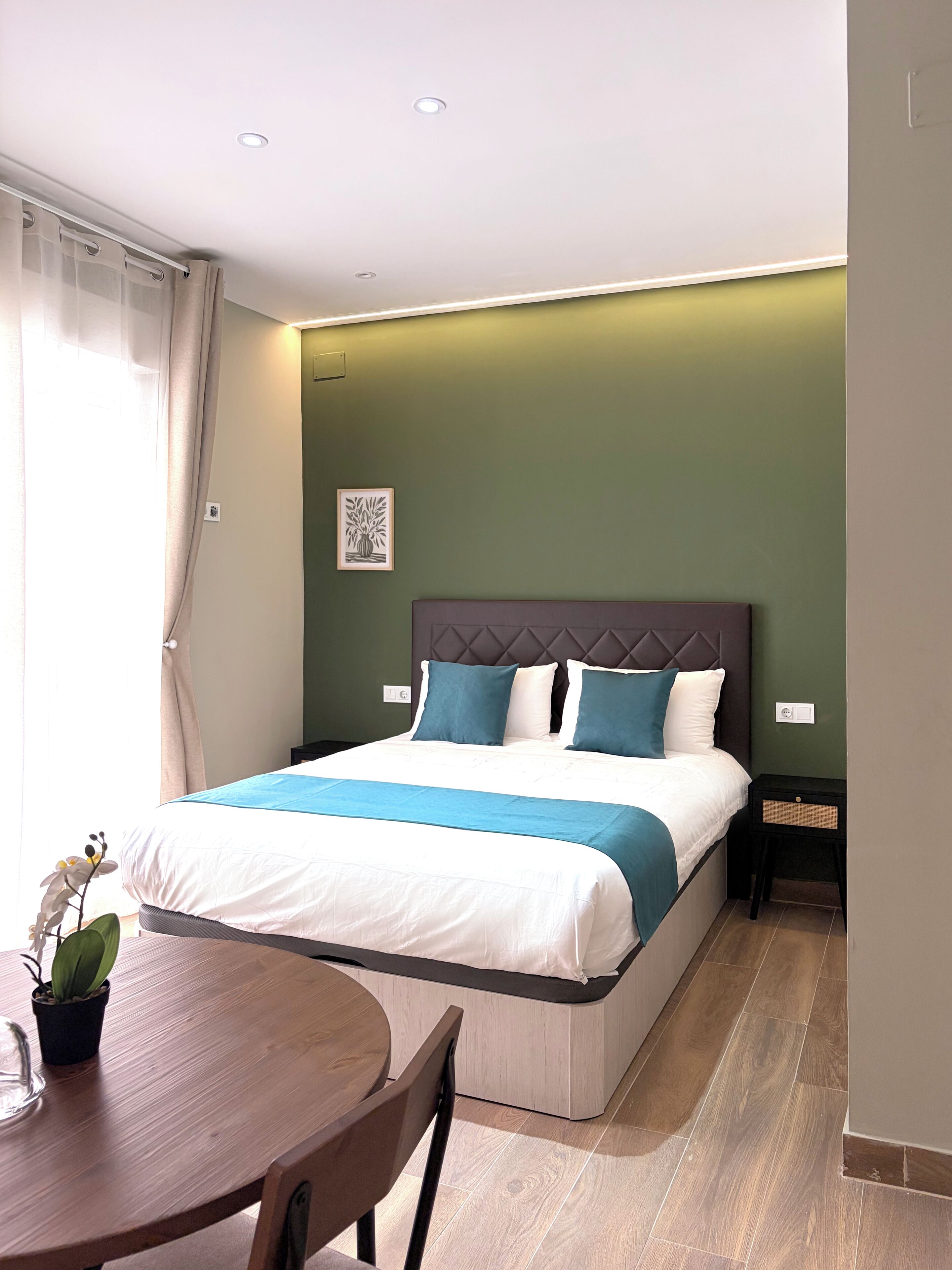 A bedroom with a bed, bedside tables, a nightstand, a chair, a table, and a potted plant.