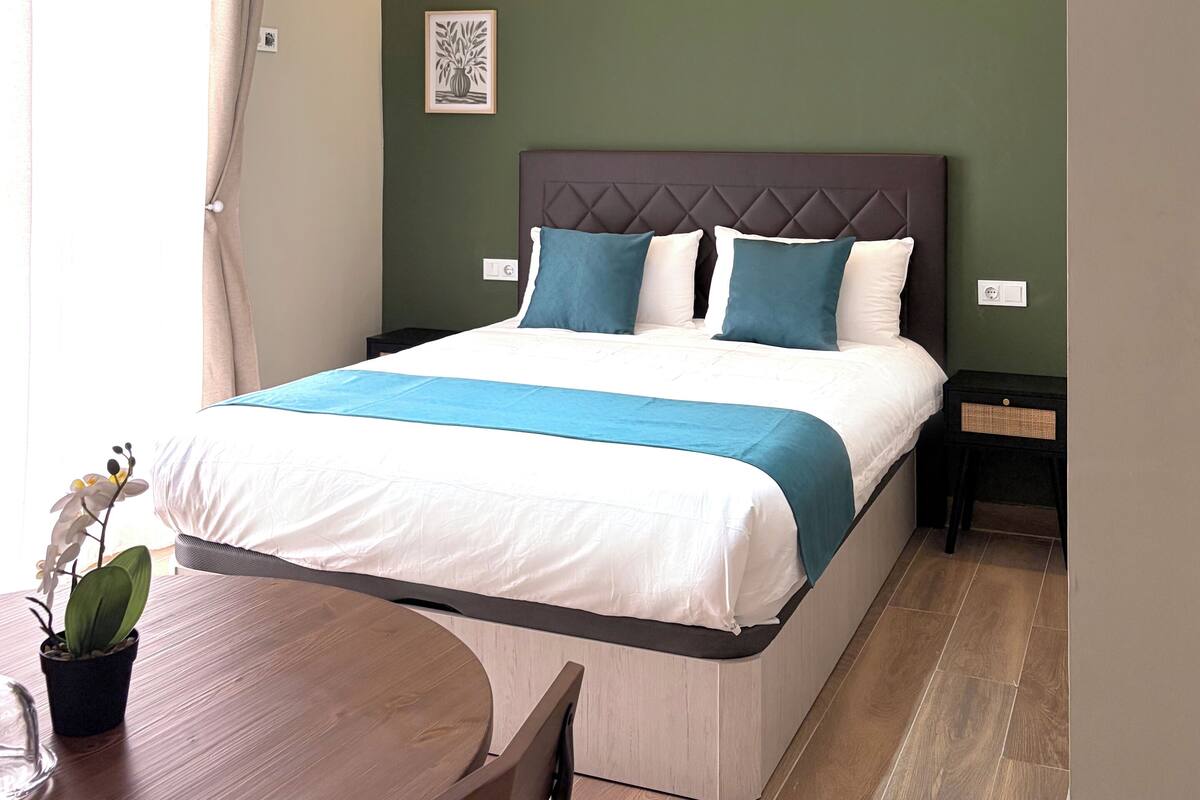 A bedroom with a bed, bedside tables, a nightstand, a chair, a table, and a potted plant.