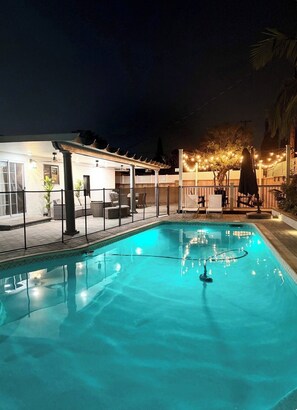 Ein Swimmingpool mit einer Pergola und einer Sitzgelegenheit im Freien, umgeben von einem Zaun und Lichterketten.