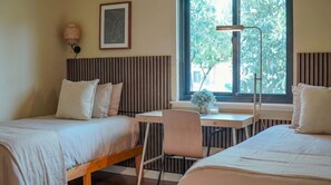 Ein Schlafzimmer mit zwei Betten, einem Schreibtisch, einem Stuhl, einer Stehlampe und einem Fenster mit Blick auf Bäume.