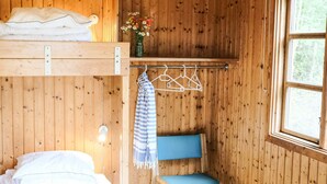 A wooden cabin interior with a bunk bed, a chair with a blue cushion, a hanging clothes rack, and a window with a view of trees. - 4 Star Holiday Home in Aakirkeby (Akirkeby)