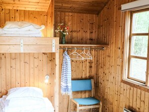 Ein Interieur einer Holzhütte mit einem Etagenbett, einem Sessel mit blauer Sitzkissen, einem Kleiderständer und einem Fenster mit Blick auf Bäume.