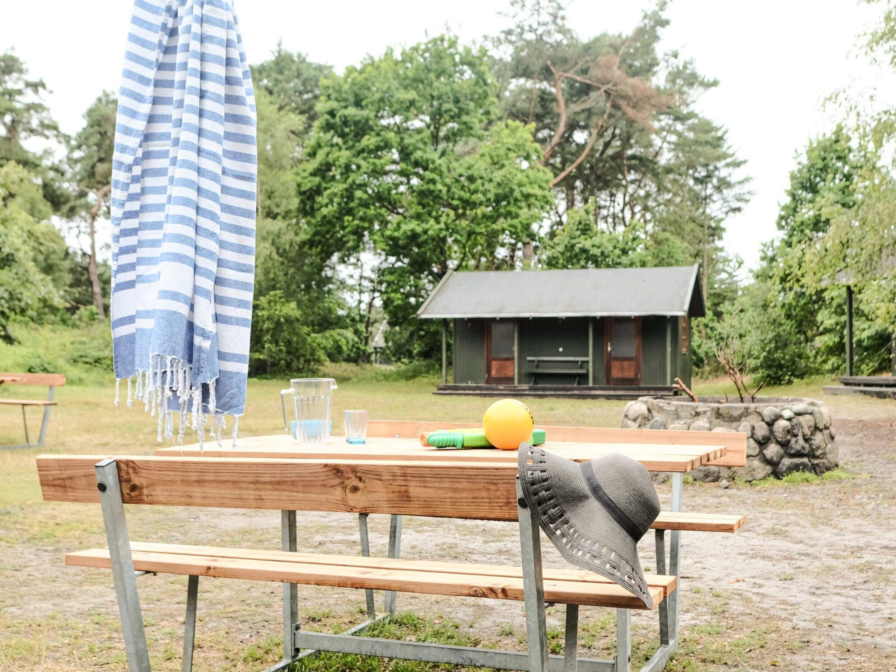 A wooden bench with a hat, a striped towel, and a yellow ball on a table in a natural setting with a cabin in the background.