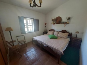 A bedroom with a bed, a chair, a table, a window, and a shelf with decorative items.