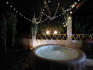 An outdoor hot tub area with string lights, surrounded by greenery and a wooden fence.