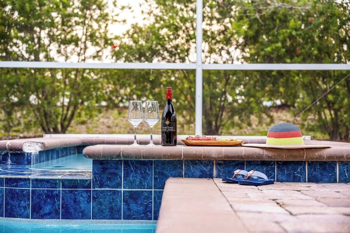 A pool area with a tiled edge, a bottle of wine, two glasses, a hat, and a pair of sunglasses.