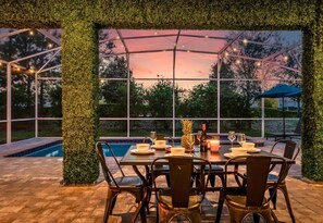 Ein Essbereich im Freien mit Tisch für vier Personen, umgeben von Stühlen, in der Nähe eines Pools mit Blick auf Bäume und einen Sonnenuntergang.