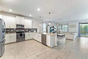 A modern kitchen with white cabinets, a built-in oven, and a dishwasher. There is a dining area with a white table and chairs. - Resort Lazy River! Themed 9BR! Close to Disney! (Four Corners)