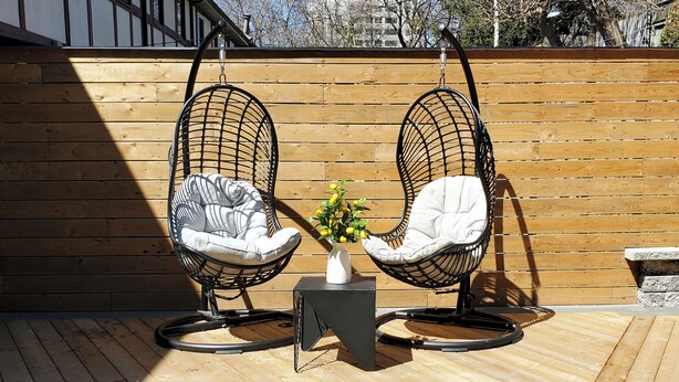 Two black wire egg chairs with cushions, a small white vase with yellow flowers, and a black square table on a wooden deck.