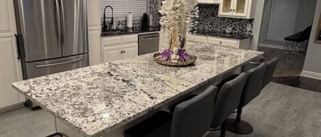 A modern kitchen with a granite island, stainless steel appliances, and a trash bin.