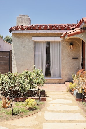 A building with a tiled roof, a window with a curtain, a pathway leading to the entrance, and various plants in the garden. - Spanish Oasis in Miracle Mile w/ Backyard (Los Angeles)