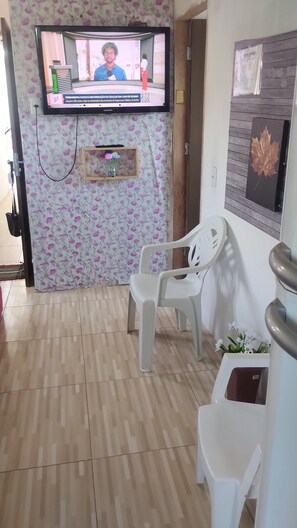 A room with a television displaying a news broadcast, a white plastic chair, a wooden shelf with a vase of flowers, and a wooden door.