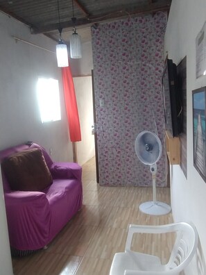 Una habitación con un sofá morado, una silla blanca, un ventilador de pie, una cortina roja y una pared con estampado floral.