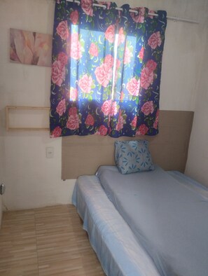 Un dormitorio con cama, almohada, cabecera, estante en la pared y ventana con cortinas florales.