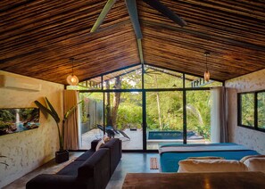 A room with a large glass wall, a black sofa, a bed, and a hanging light fixture. - Pond House l Waterfront Jungle Retreat w/Private Pool, Fire Pits, Hammocks & AC (Provincia de Puntarenas)
