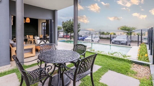 Une terrasse avec une table ronde et des chaises, une piscine, et une vue sur les voitures garées et les bâtiments.