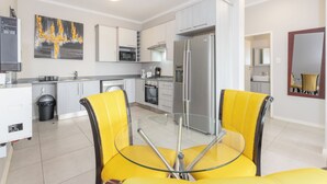 Une cuisine moderne avec des meubles blancs, un réfrigérateur en acier inoxydable et une table à manger en verre, assortie de chaises jaunes.