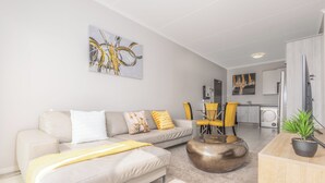 Un salon moderne avec un canapé, une table basse et des fauteuils. On aperçoit un coin cuisine au fond.