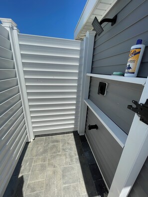 Un petit espace extérieur avec une porte coulissante blanche, un sol carrelé gris et une étagère sur laquelle se trouvent un flacon de shampoing et un porte-savon.
