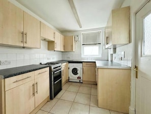 A compact kitchen with light wood cabinets, a built-in oven, a washing machine, and a window with blinds. - 3 BR House | Sleeps 7 | Free Parking | Fast Wi-Fi (Salford)