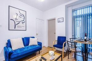 Ein modernes Wohnzimmer mit einem blauen Sofa, einem Esstisch mit Stühlen, einem gerahmten Kunstwerk an der Wand und einer Tür zu einem weiteren Raum.