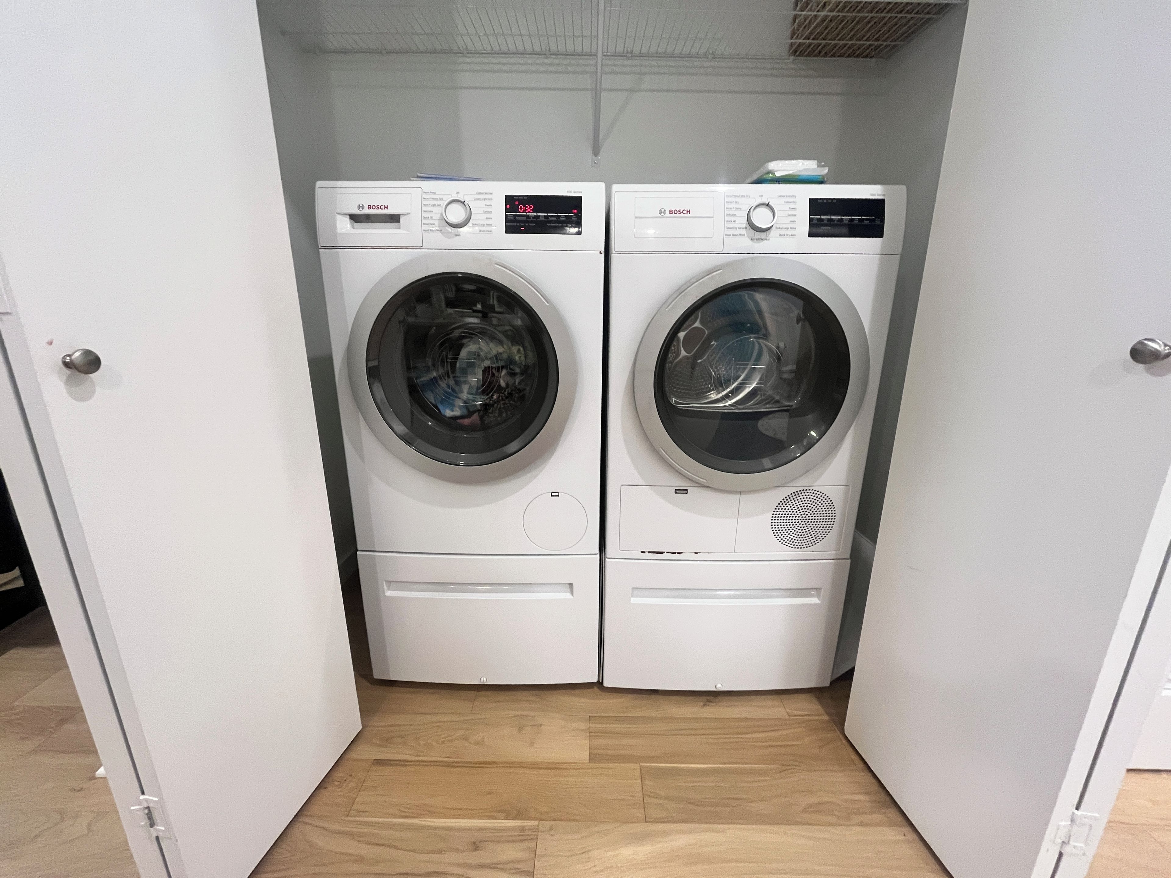 A compact laundry room with two white Bosch washing machines, a wooden floor, and a white door.
