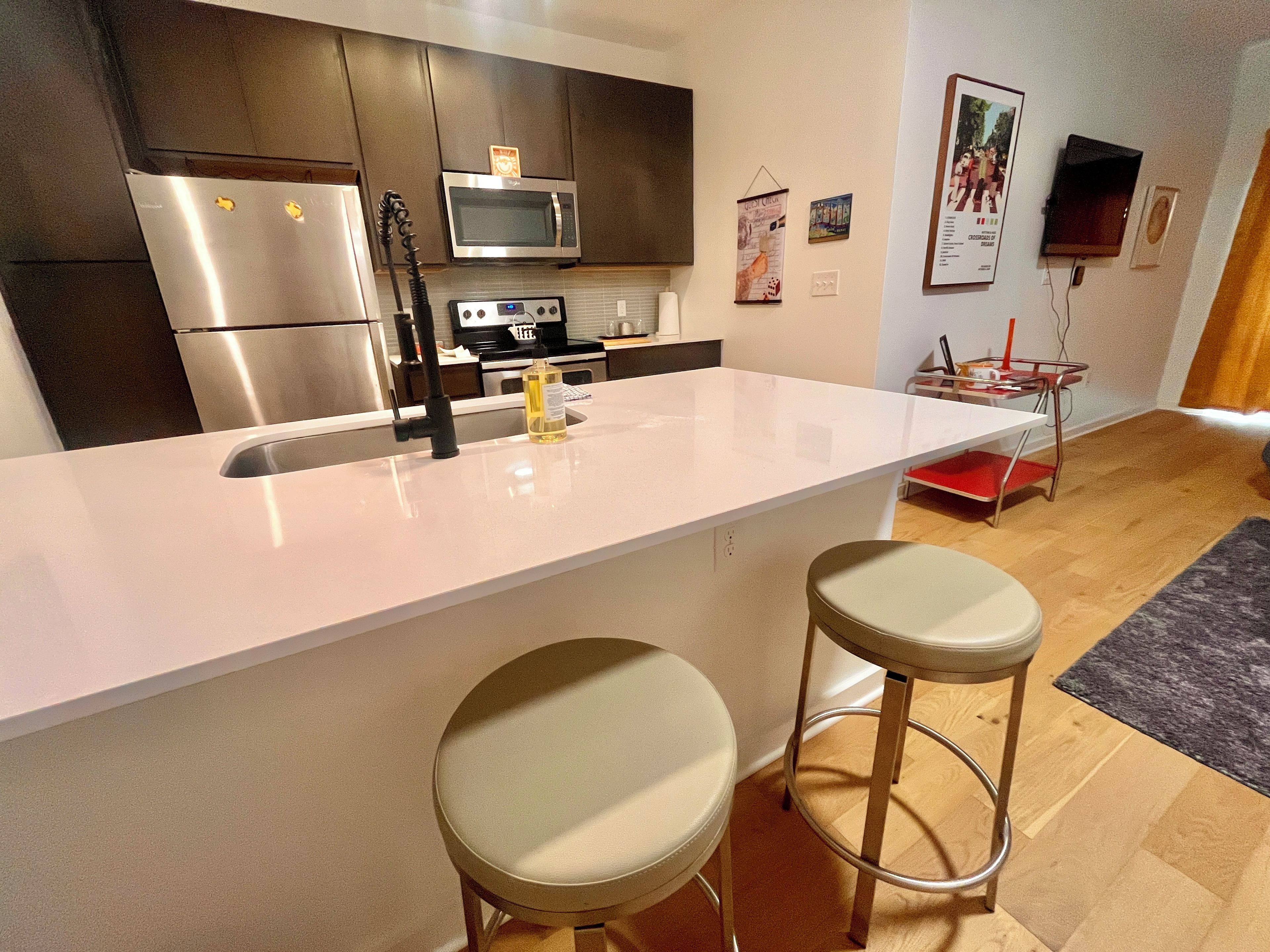 A modern kitchen with stainless steel appliances, a white countertop, and two bar stools.
