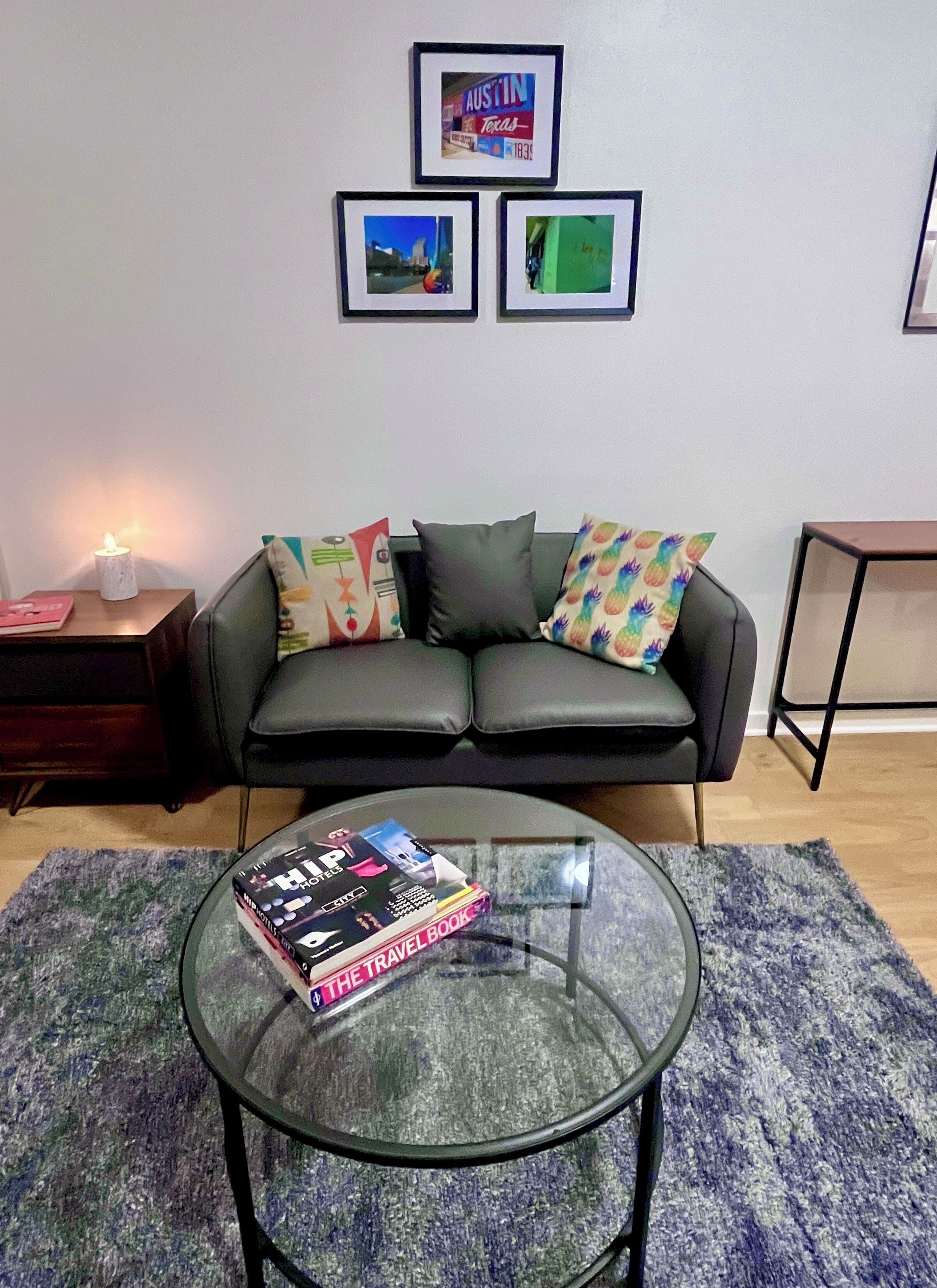 A living room with a black leather sofa, a glass coffee table with a travel book, and a wall with framed pictures.