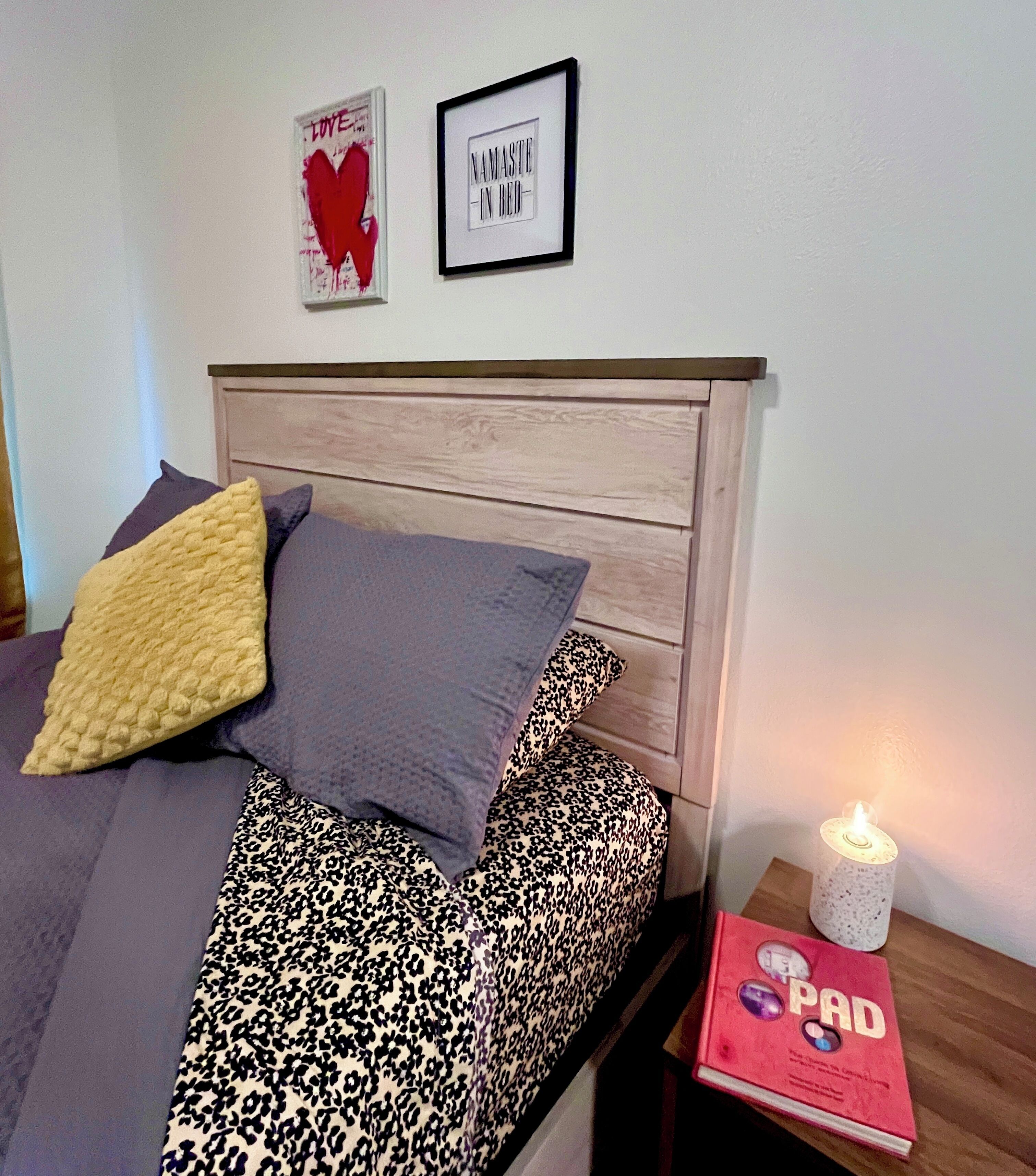 A bedroom with a bed, pillows, a bedside table with a book and a candle, and two framed pictures on the wall.