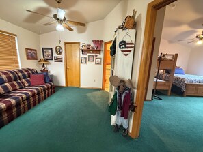 A room with a plaid sofa, a ceiling fan, a bed with a blue blanket, and a wall with framed pictures and a wooden shelf. - Grandmas Cabin a Cozy Mountain Retreat (Overgaard)