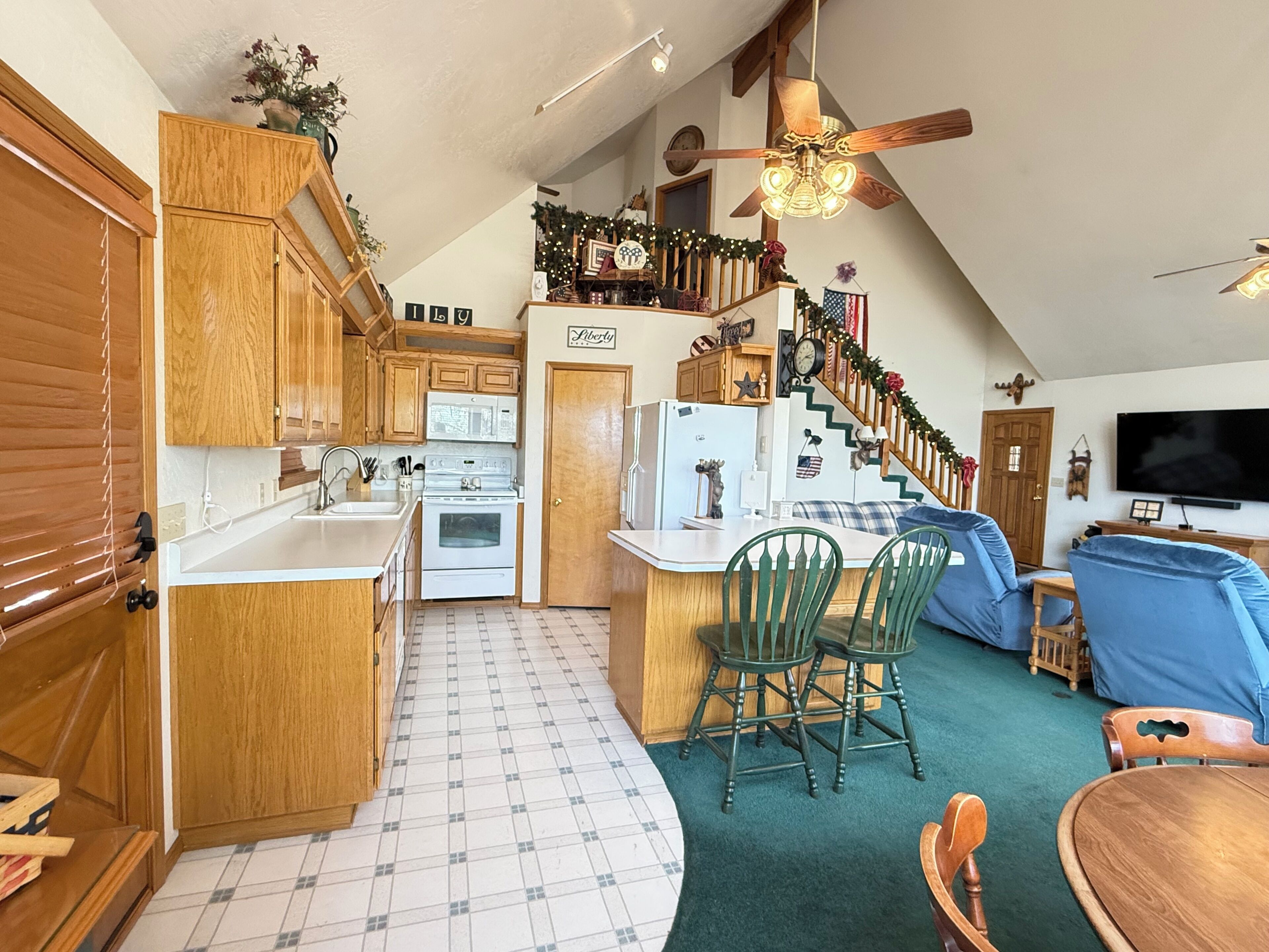A kitchen with wooden cabinets, a microwave, and a stove. There is a dining area with chairs and a table. A staircase leads to a loft with a ceiling fan.