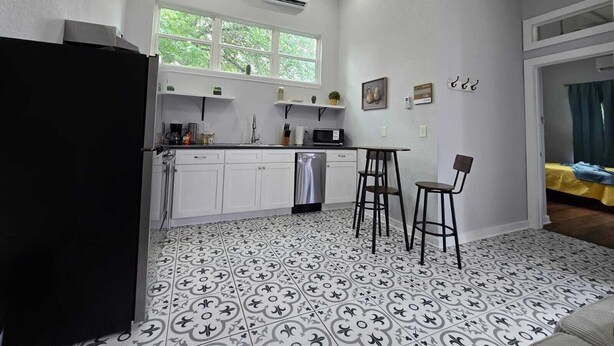 Eine kompakte Küche mit weißen Schränken, einem schwarzen Kühlschrank, einer Mikrowelle und einem Spülbecken. Zwei Barhocker und ein Fenster über dem Spülbecken.