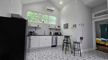 A compact kitchen with white cabinets, a black refrigerator, a microwave, and a sink. There are two bar stools and a window above the sink.
