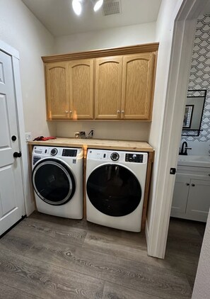 A compact laundry room with two front-loading washing machines, wooden cabinets above, and a tiled floor. - 3-bedroom pet-friendly house in Grover Beach - 2 miles from the Beach (Grover Beach)