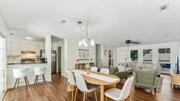 A modern kitchen with white cabinets, a dining area with a wooden table and white chairs, and a living room with a green sofa and a TV.