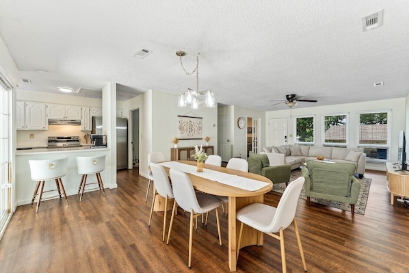 Une cuisine moderne avec des meubles blancs, un coin repas avec une table en bois et des chaises blanches, et un salon avec un canapé vert et un téléviseur.