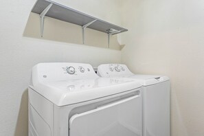 Ein Waschbereich mit zwei übereinander gestapelten Waschmaschinen und Trocknern, einem Metallregal darüber und einer schlichten Wand.