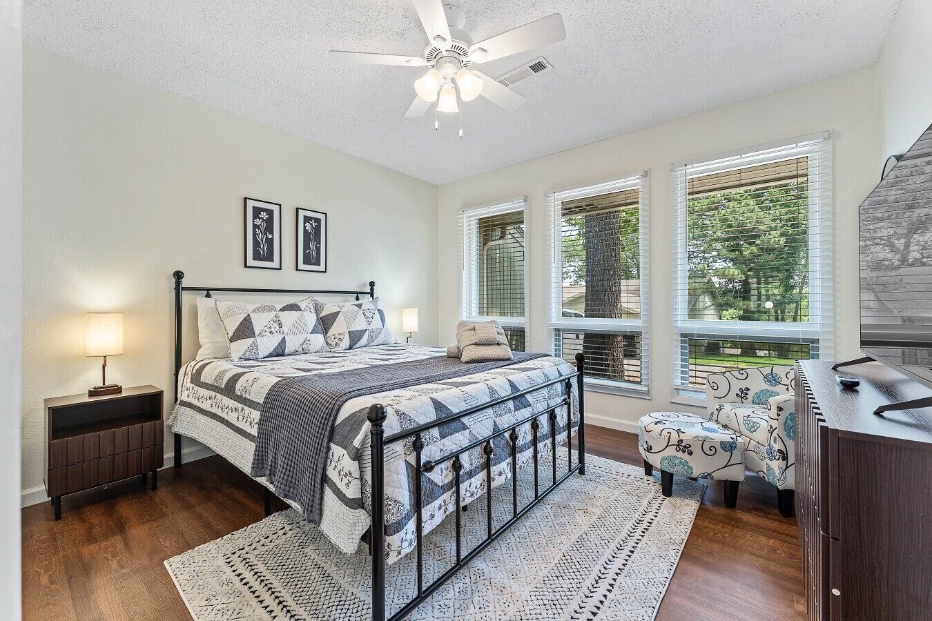 A bedroom with a bed, a desk, a chair, a ceiling fan, and large windows with blinds.