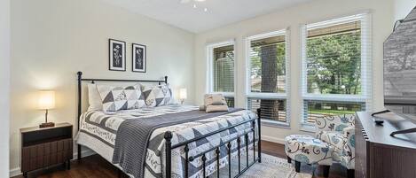 A bedroom with a bed, a desk, a chair, a ceiling fan, and large windows with blinds.