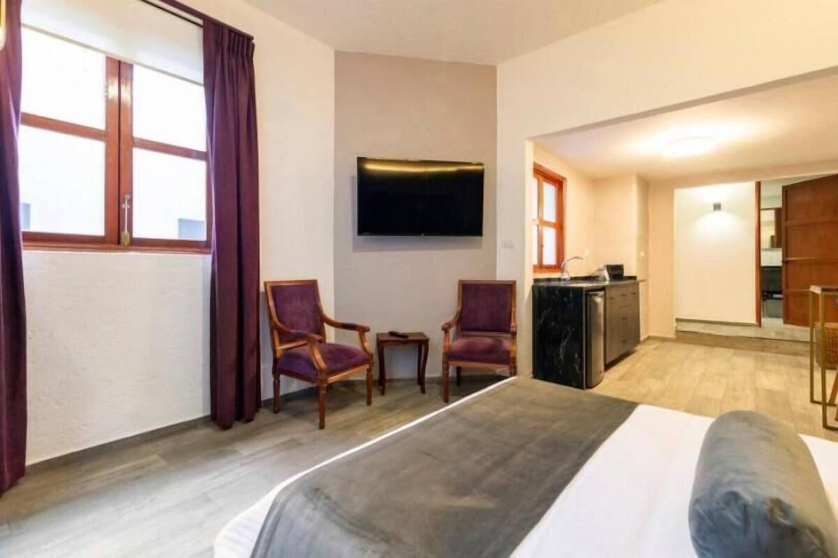 A hotel room with a bed, two armchairs, a TV, a desk with a sink, and a view of the outside.