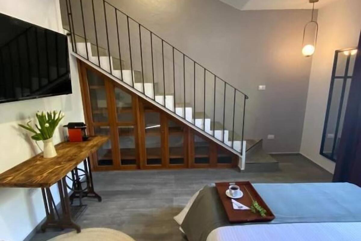 A room with a bed, a desk, a chair, a staircase, and a hanging light fixture.